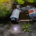 Vista aérea de casas ribeirinhas sobre área alagada, cercadas pelas águas e pela vegetação amazônica.