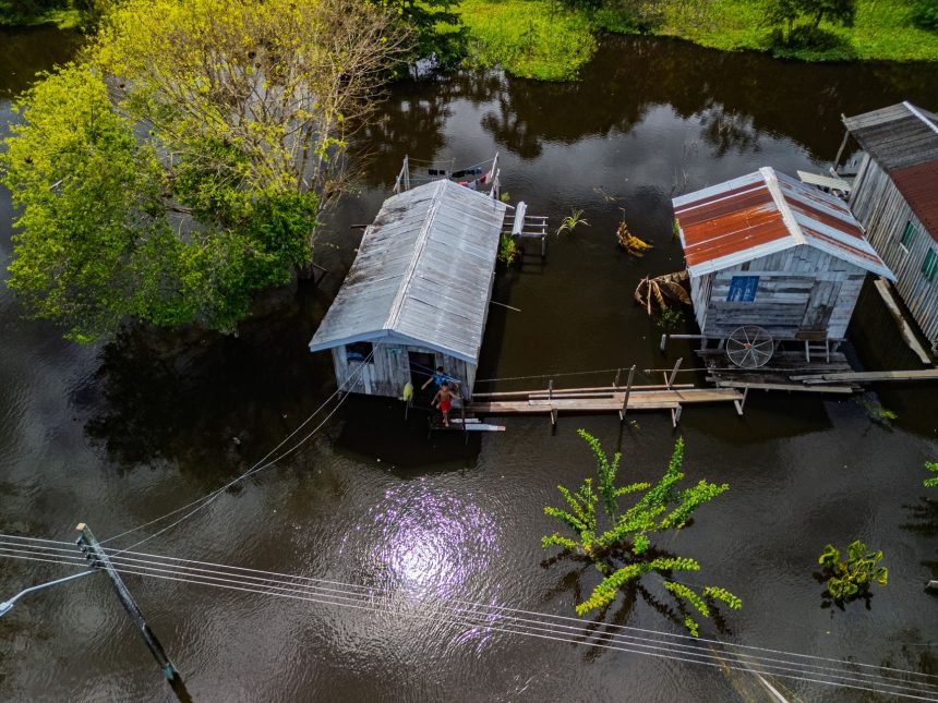 Vista aérea de casas ribeirinhas sobre área alagada, cercadas pelas águas e pela vegetação amazônica.