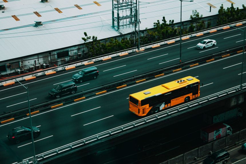 Ônibus urbano amarelo circula por uma avenida movimentada, visto de cima, entre outros veículos.